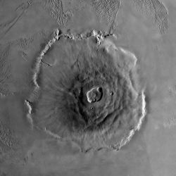 Olympus Mons, a shield volcano, is the tallest volcano in the solar system. It rises 27 kilometers (17 miles) high, spreads over 600 km (370 mi) at the base, and is surrounded by a well-defined scarp that is up to 6 km (4 mi) high. Lava flows drape over the scarp in places. Much of the plains around the volcano are covered by ridged and grooved terrain called the aureole. The origin of the aureole is controversial, but may be related to material sliding off of the flanks of an ancestral volcano. The summit 
